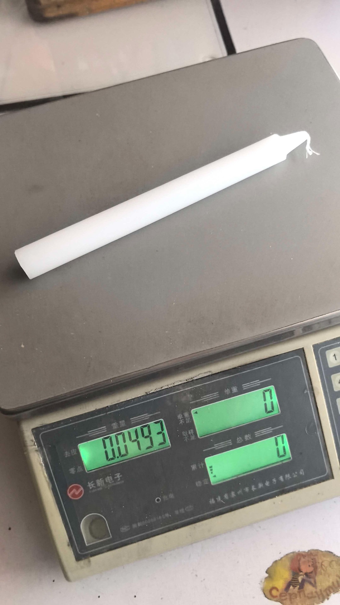 White taper candle on a scale showing a measured weight of 0.04493 kilograms, equivalent to 44.93 grams